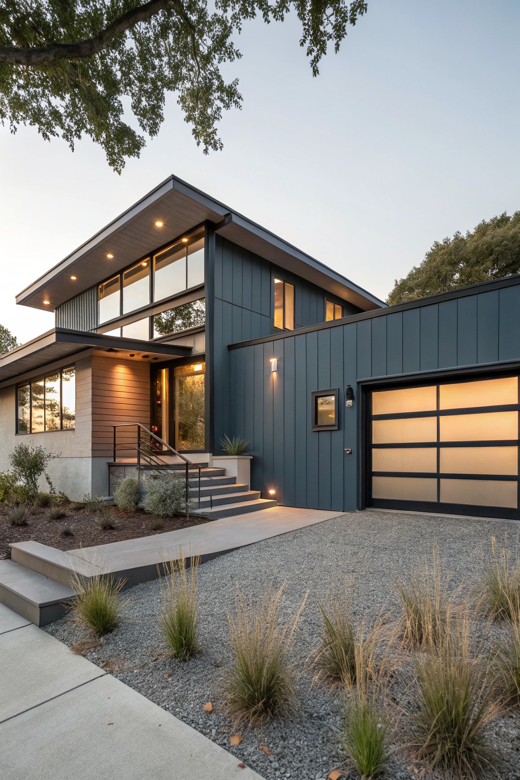Modern two-story house with dark blue vertical siding, warm wood accents on the entryway and upper levels, frosted glass garage door, concrete steps, and gravel yard planted with ornamental grasses.