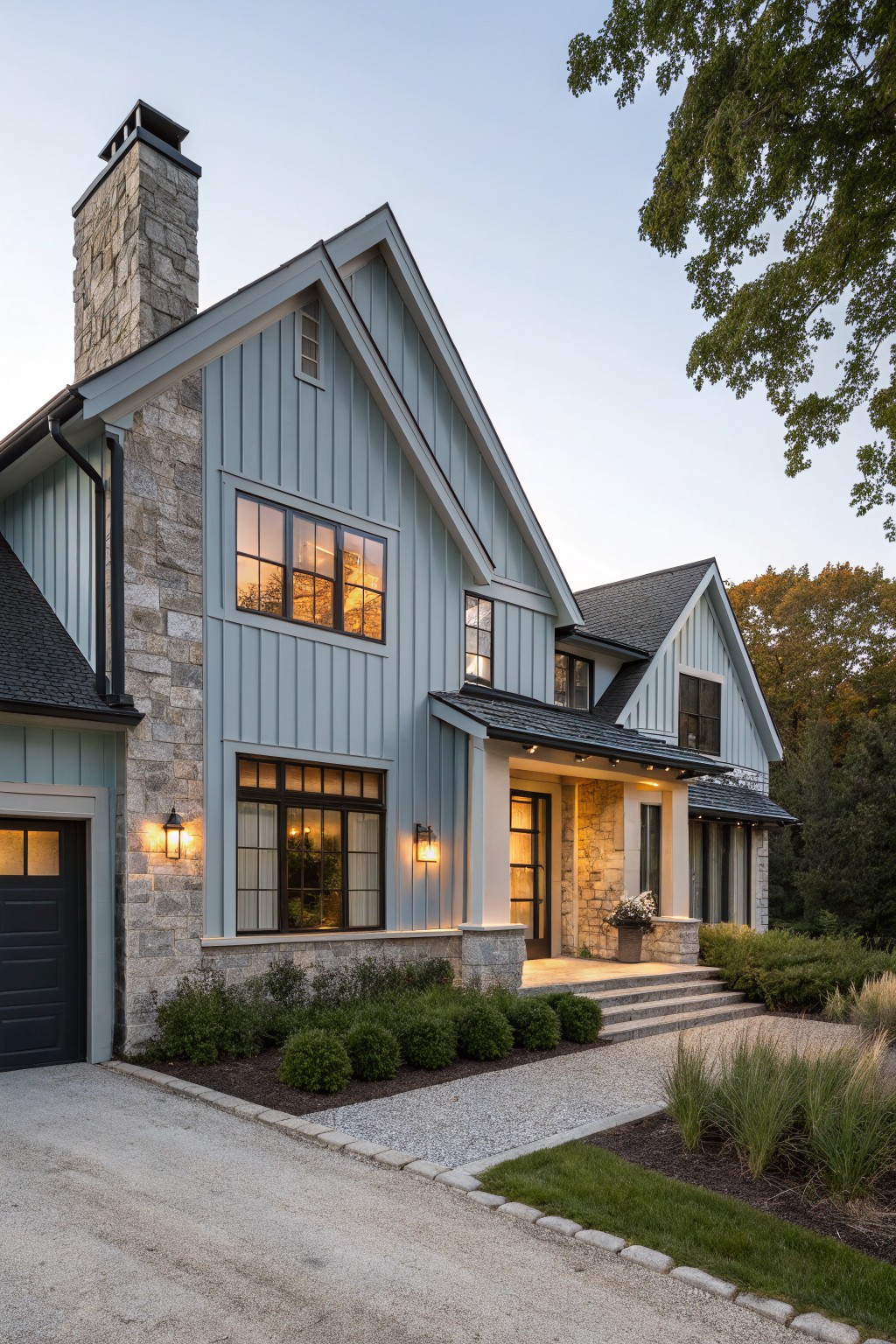 Two-story blue clapboard house with stone veneer on chimney, garage, and entry porch, large windows, black roof, landscaping with shrubs and gravel path at dusk.