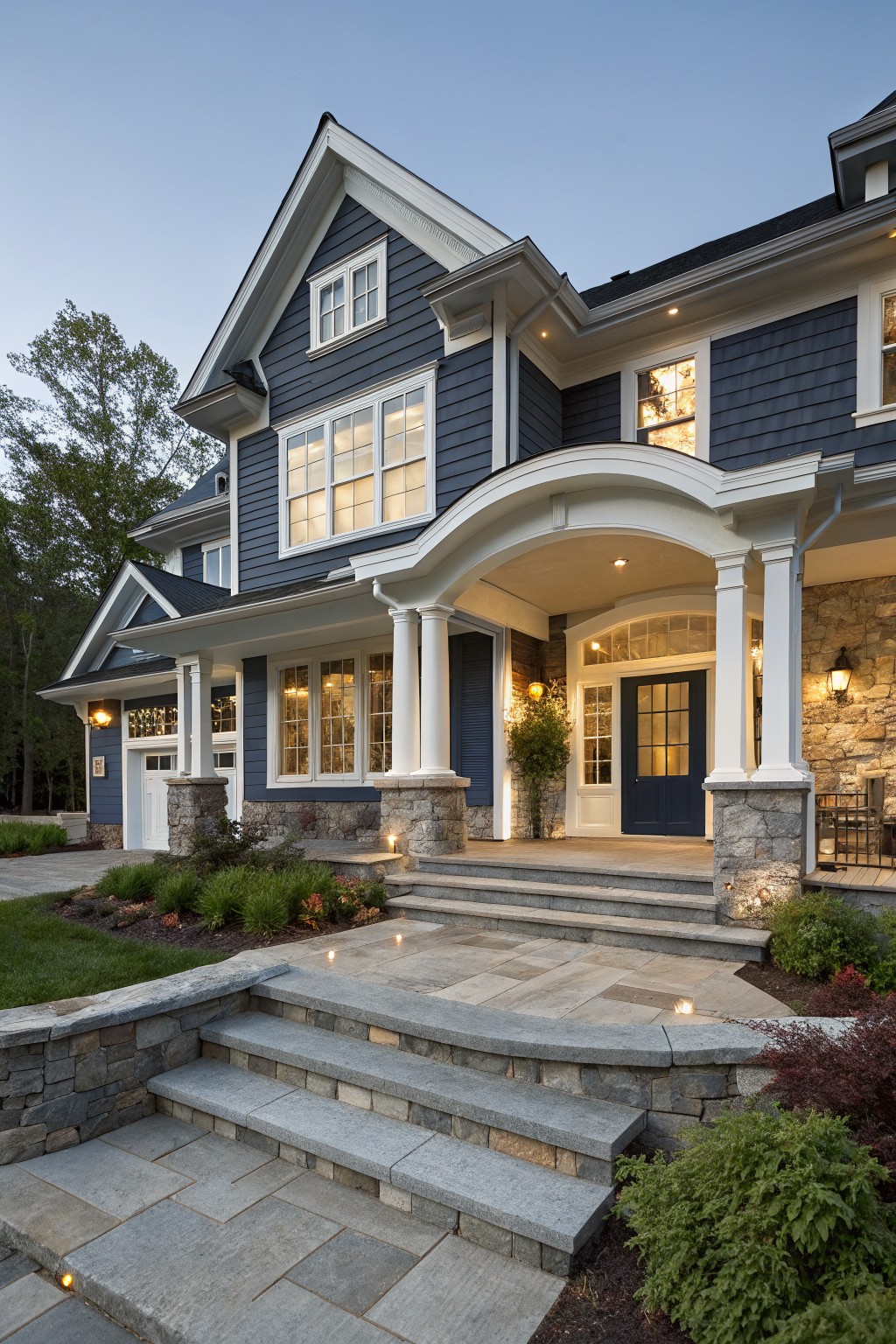 Two-story blue shingle-style house exterior with white trim, stone veneer on porch pillars, garage foundation, and retaining walls, plus steps leading to an arched entryway and landscaping.