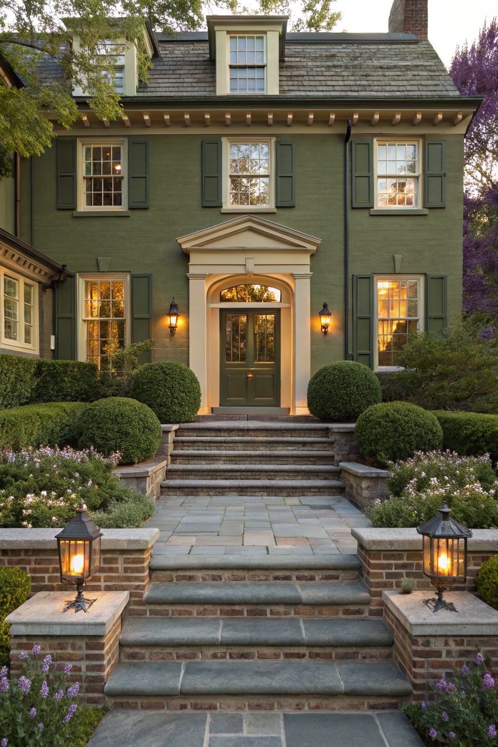Green painted house exterior with symmetrical stone steps leading to a centered front door, flanked by spherical boxwood shrubs, pedestal lanterns, and low flower beds.