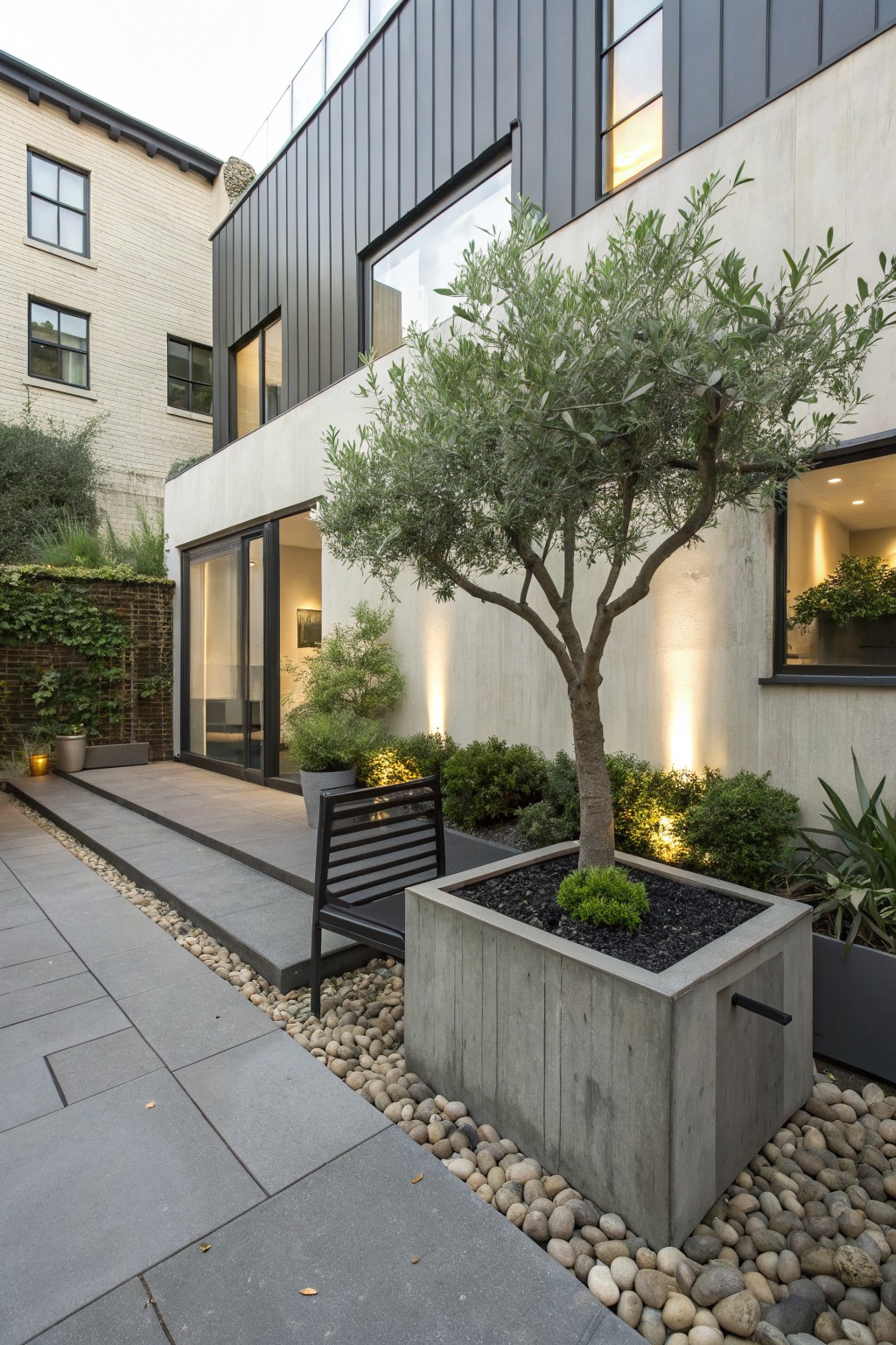 Modern backyard patio with dark slate tiles, white pebble mulch borders, central olive tree, concrete planter with small shrub, black metal bench, steps, and sliding glass doors on a light concrete wall with uplights.