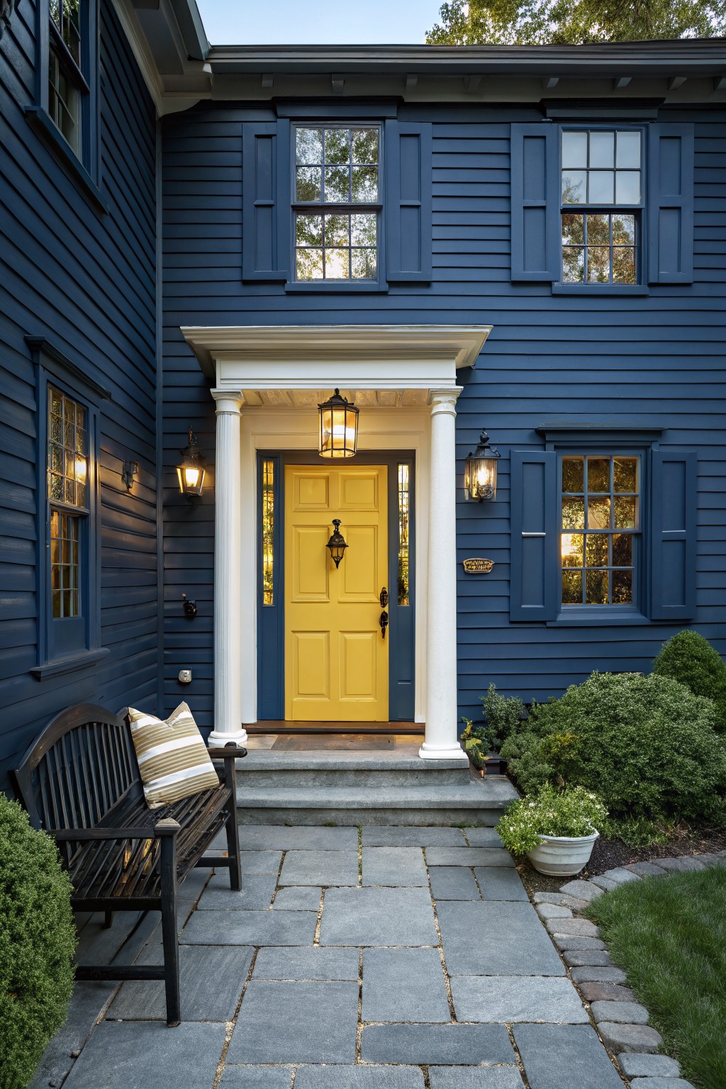 Navy blue clapboard house exterior featuring a bright yellow paneled front door under a white portico with columns and lanterns, flanked by windows with shutters, a stone pathway, black bench, and shrubs.