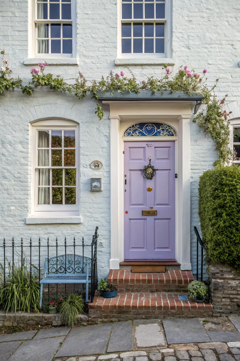 White brick house exterior with lavender purple paneled front door, brass hardware, climbing pink roses, blue bench, and potted plants on a stone path.