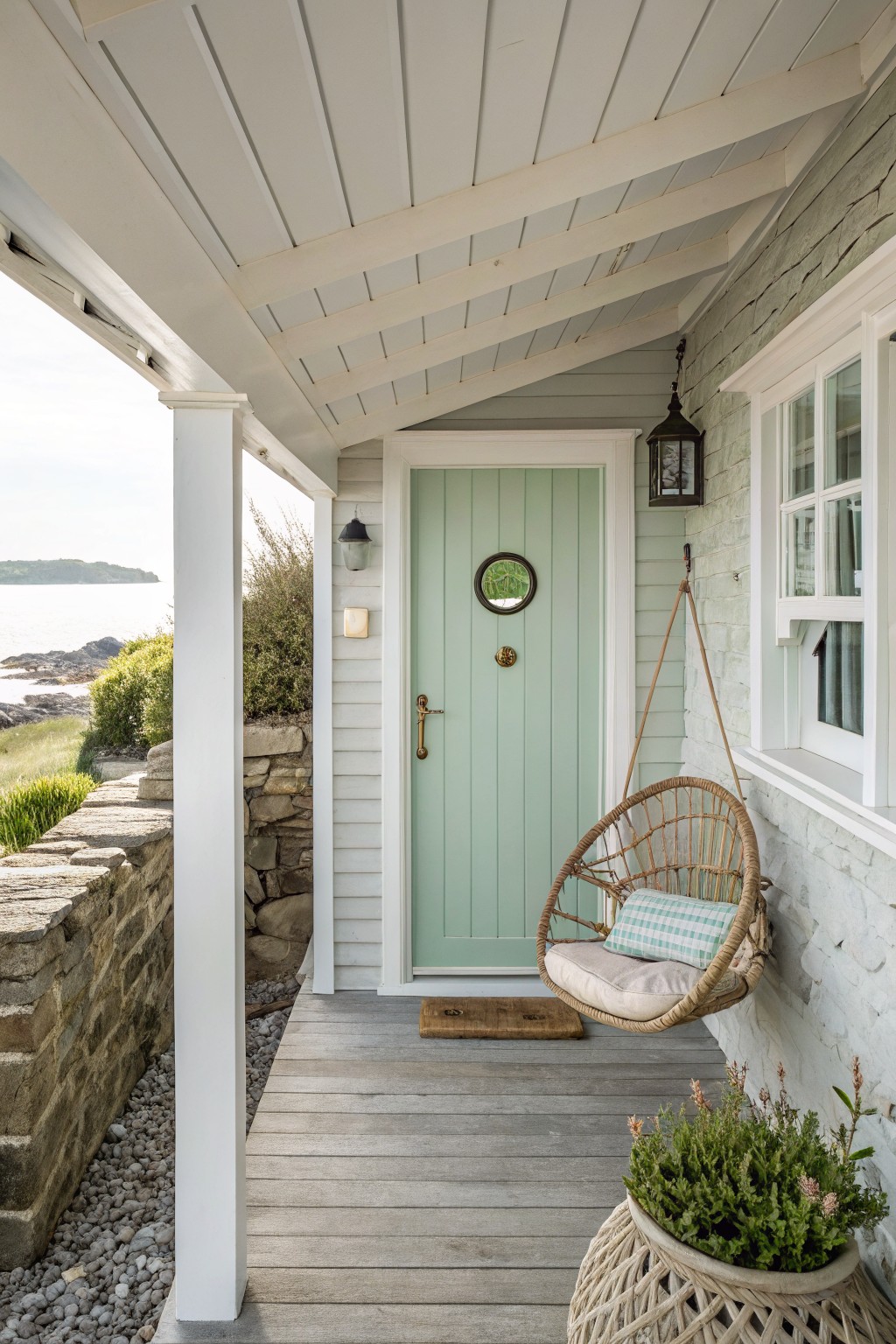 Mint green paneled front door with porthole window and brass knocker on a light blue shingle house exterior, featuring a hanging wicker porch swing, lanterns, stone wall, and plants on a wooden porch with sea view.