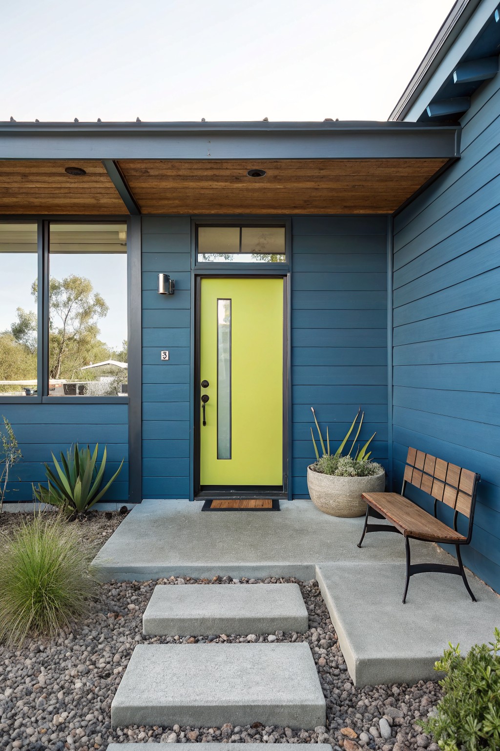 Modern house exterior painted navy blue with a bright yellow front door, large windows, wood ceiling overhang, potted agave plants, wooden bench, and concrete steps amid gravel landscaping.