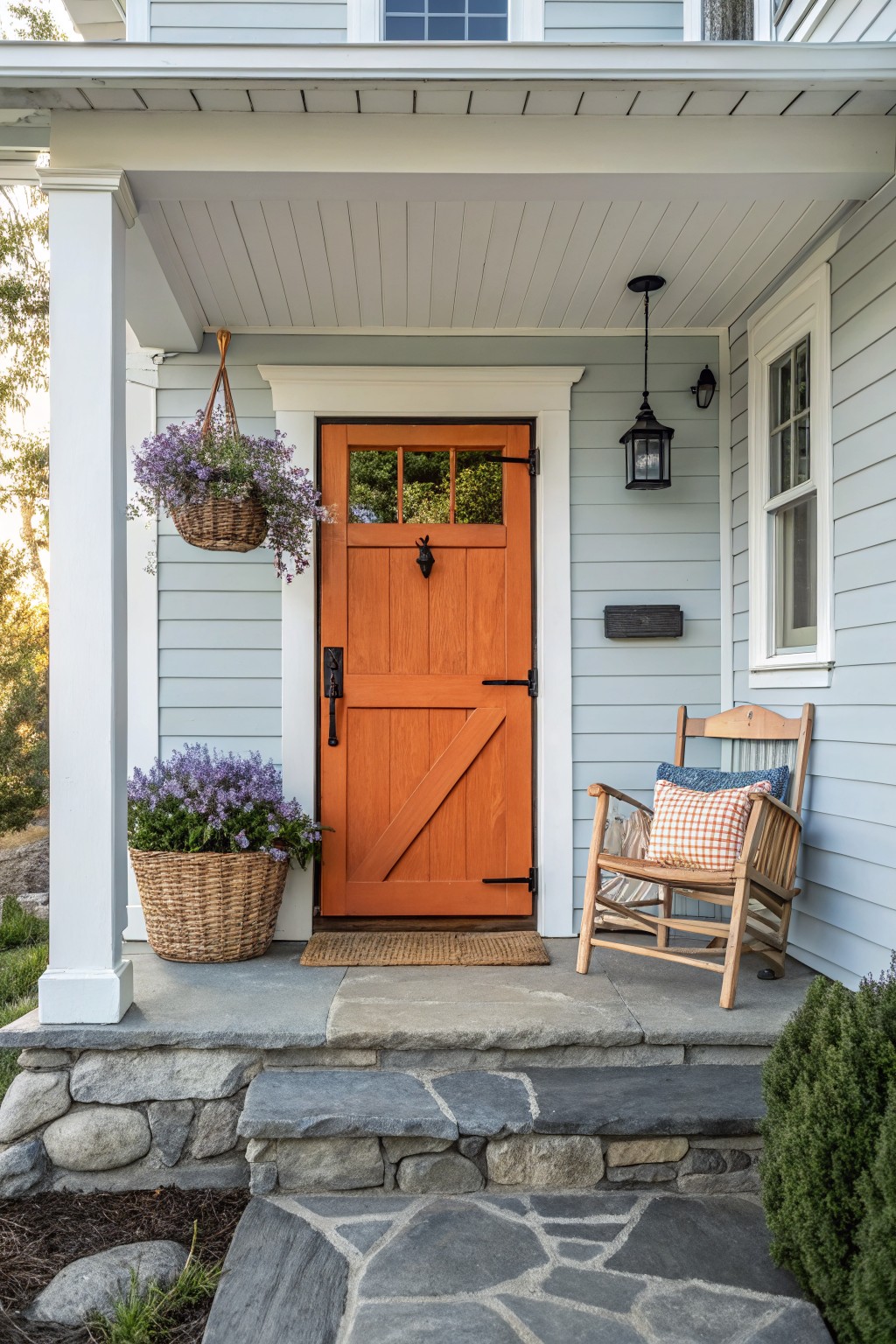 Light blue clapboard house front porch with orange wooden paneled door featuring glass inserts, white columns, hanging basket plants, a wooden rocking chair with cushions, lanterns, doormat, and stone steps.