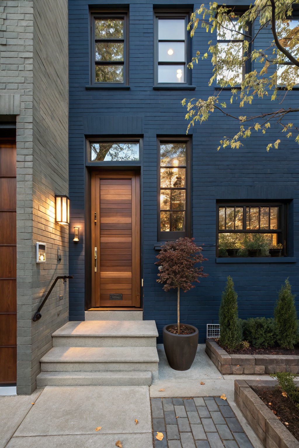 Navy blue brick house exterior with tall wooden front door featuring vertical slats, flanked by wall lanterns, concrete steps, potted Japanese maple tree, and low shrubs in raised beds.