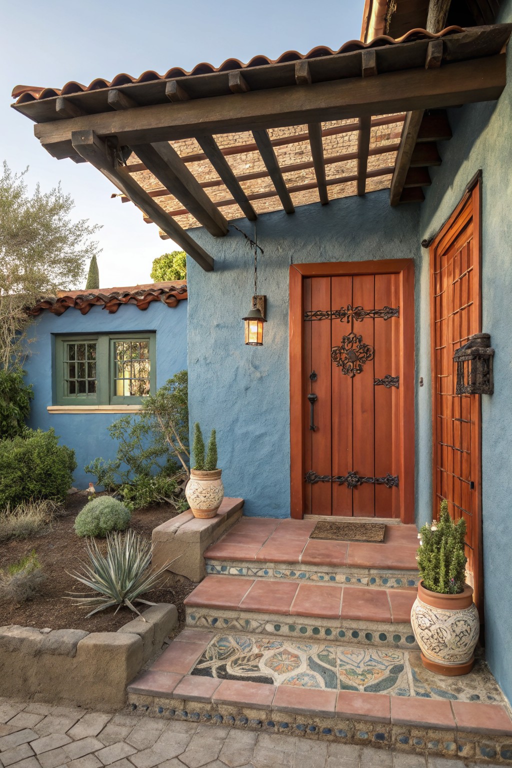 Blue stucco house entrance with a tall wooden door featuring black iron hardware and knocker, terracotta steps with tile accents, potted succulents, and a wooden pergola overhang.