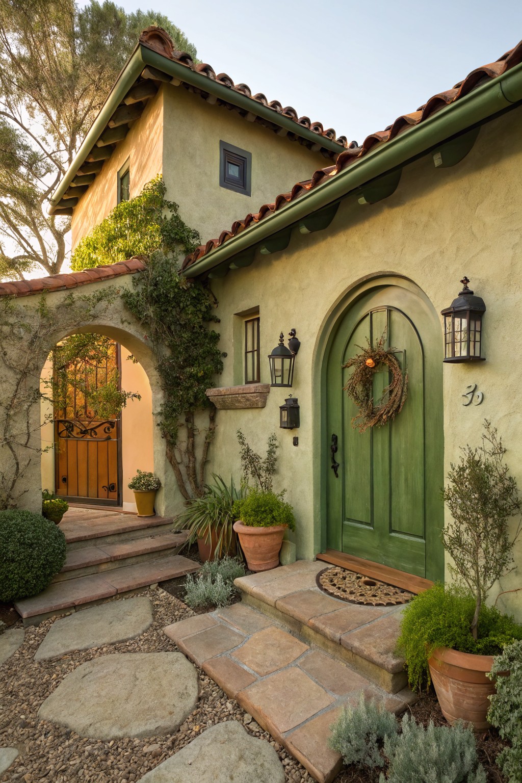 Beige stucco house exterior with red tile roof, arched green wooden front door decorated with a dried fall wreath, arched gateway with orange inner door, stone steps, potted plants, and lanterns.