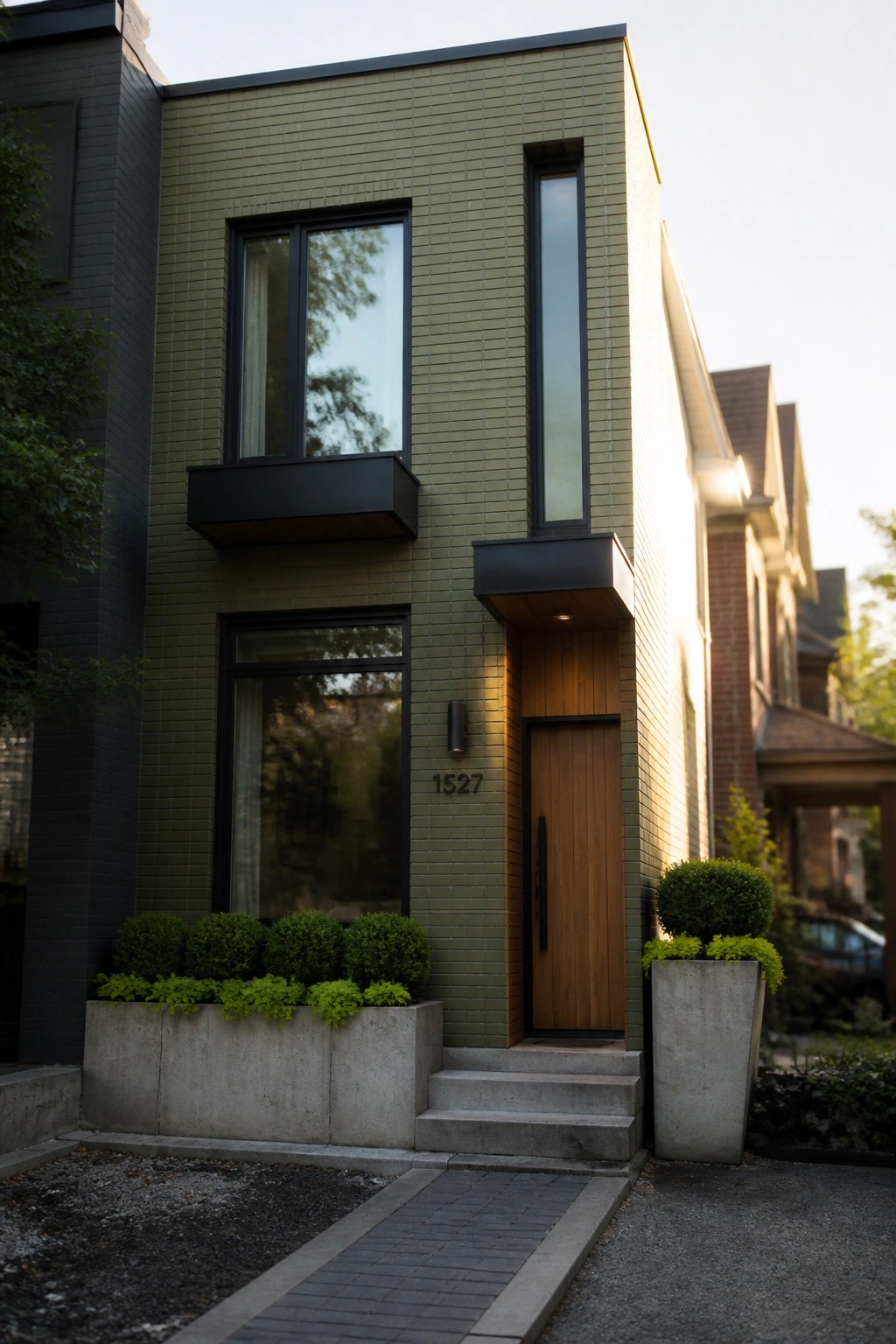 Modern two-story house with dark green brick exterior, vertical window, wooden front door, black accents, and concrete planters with shrubs at the entry.