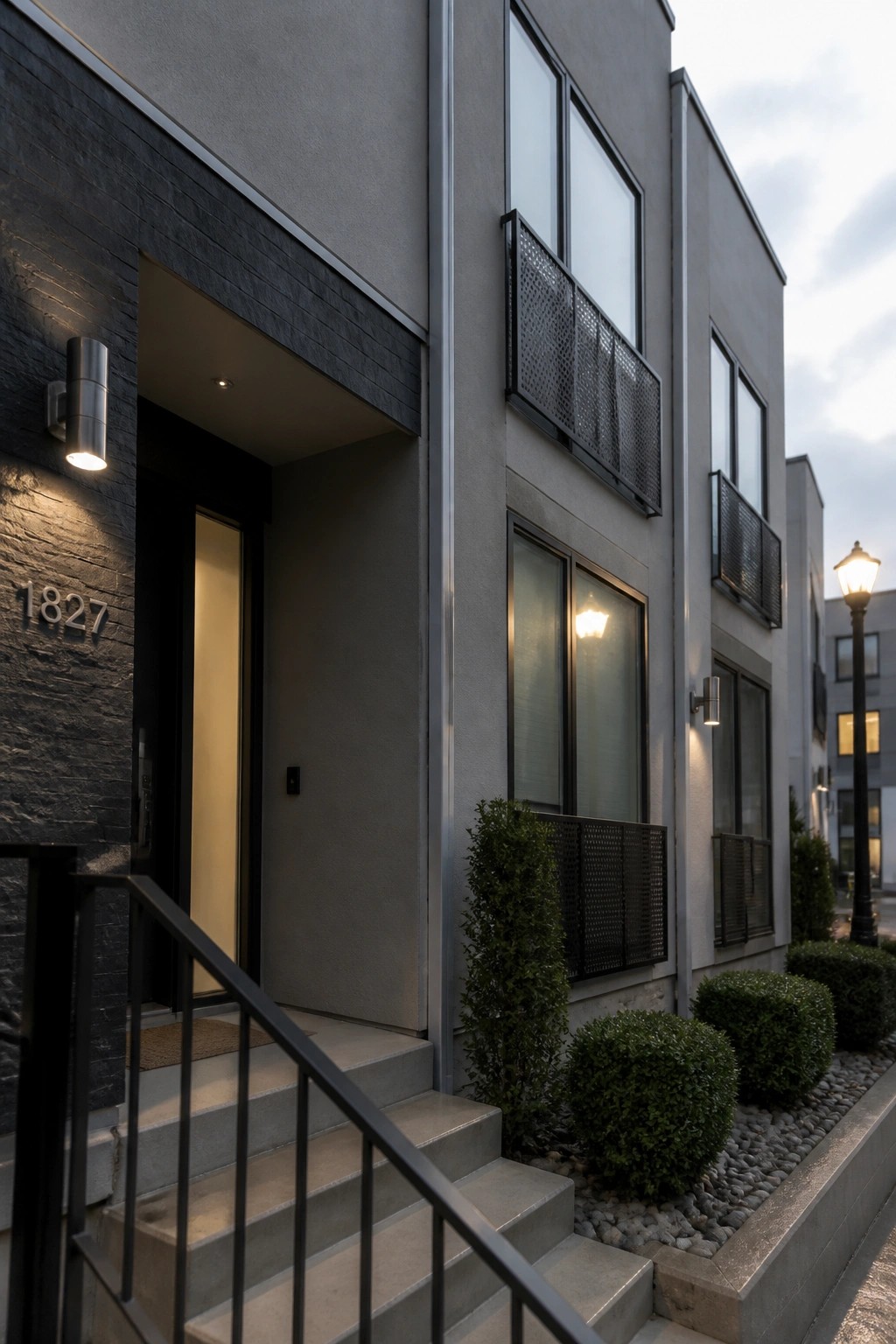 Modern gray house exterior with dark stone cladding around the front door, concrete steps, black metal railings, and a small balcony.