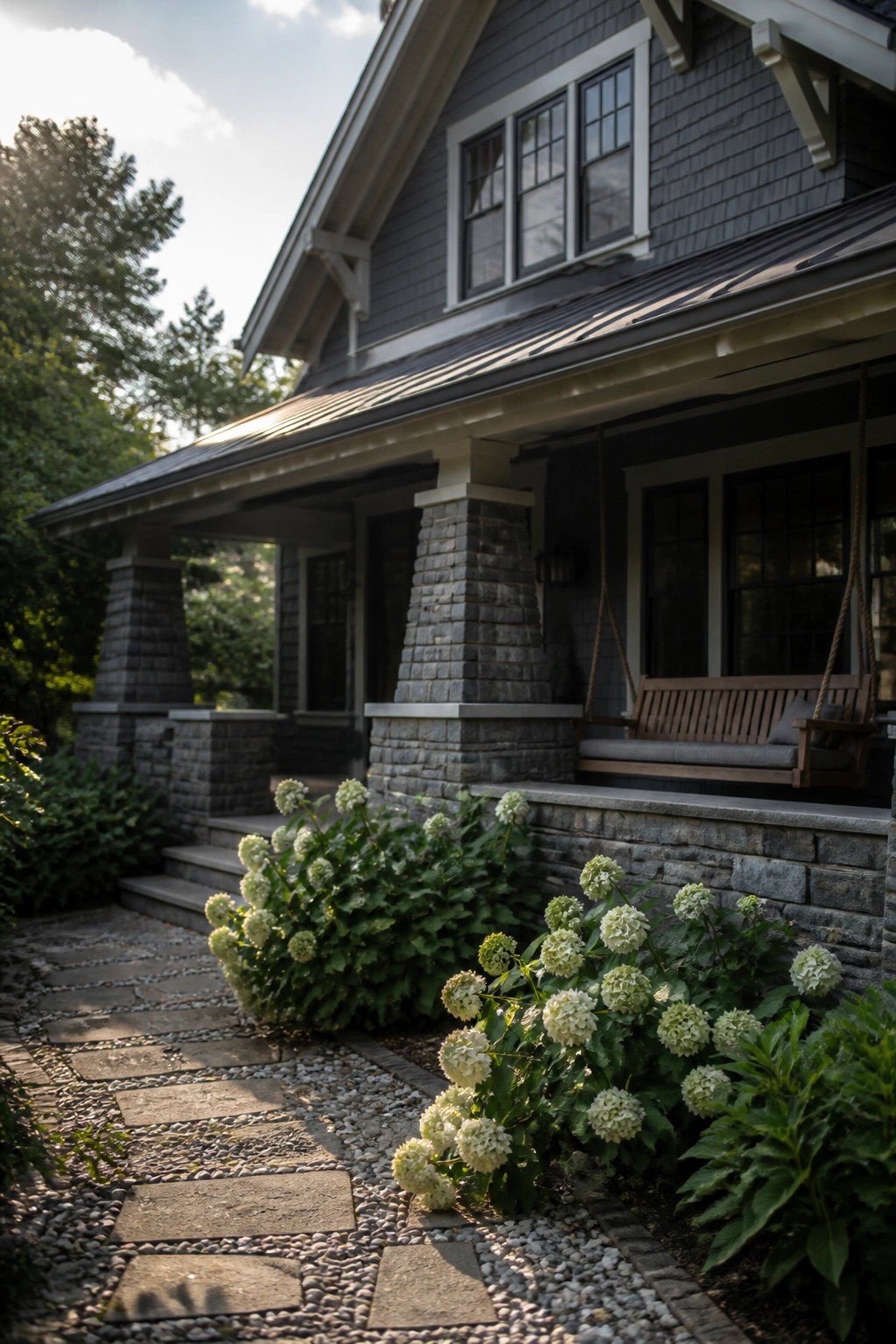 A gray shingled house with stone porch columns, a metal roof, a wooden porch swing, and white hydrangeas beside a stone pathway.