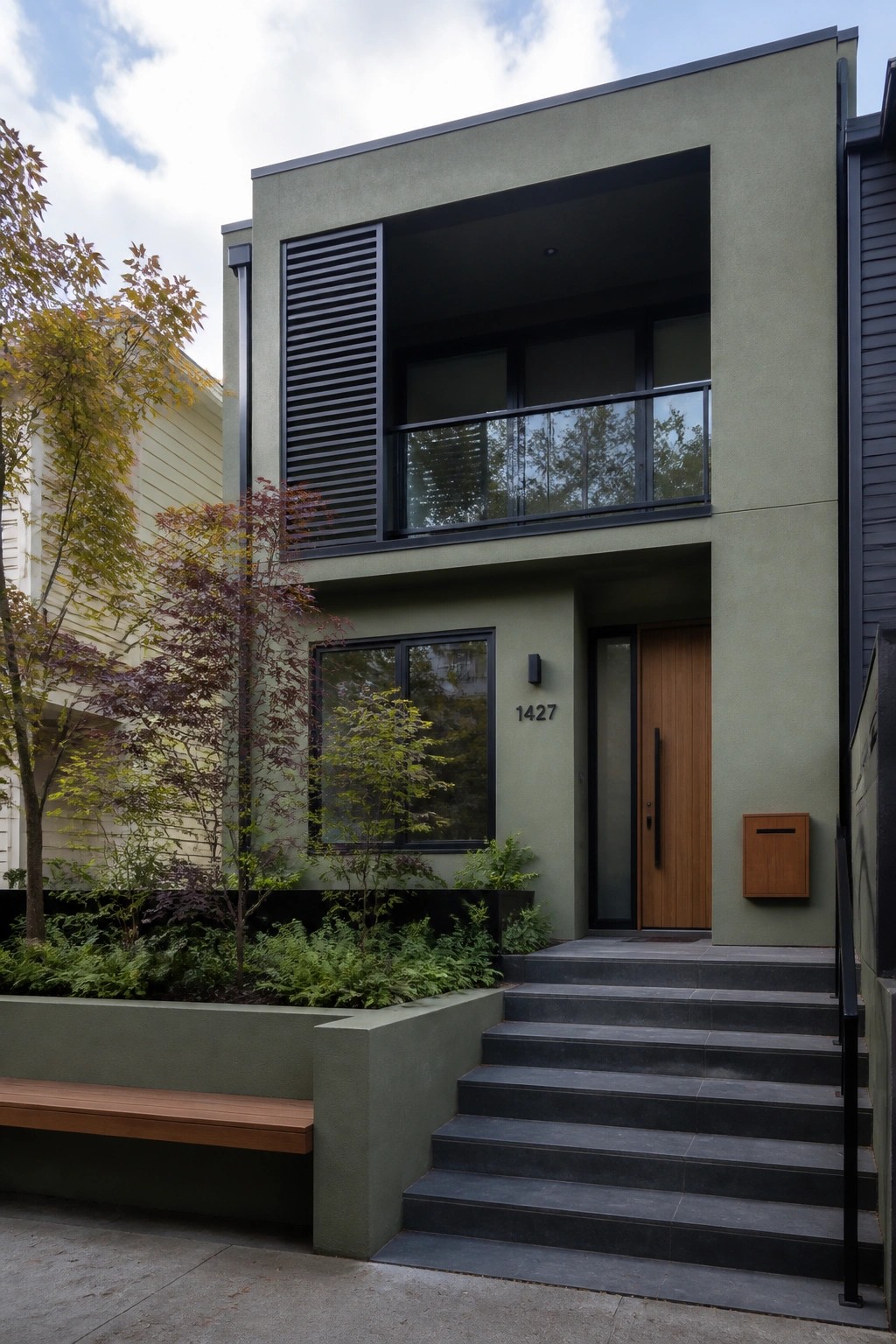 Modern two-story house with sage green stucco exterior, black-framed windows and balcony, wooden front door, and dark stone steps.