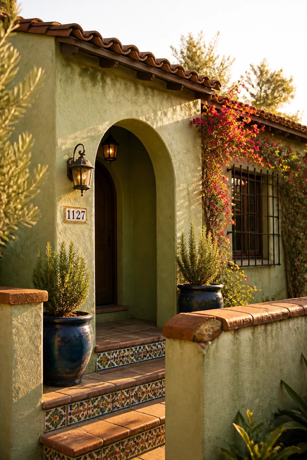 A sage green stucco house exterior with an arched doorway, terracotta tile roof, decorative wall tiles on the steps, and potted plants near the entrance.