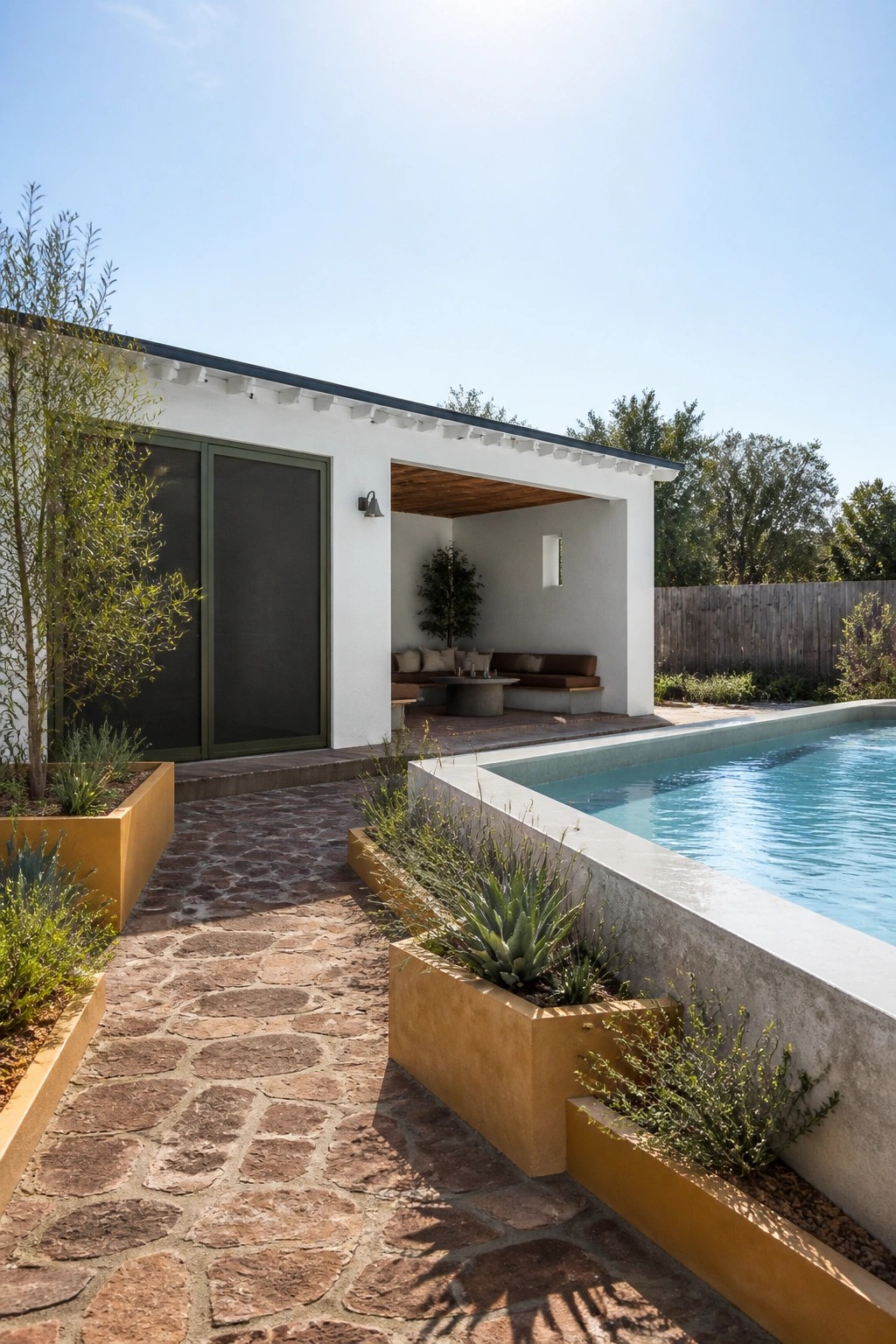 A white house exterior with olive green sliding doors, ochre planters along a stone path, built-in seating under a covered patio, and a swimming pool.