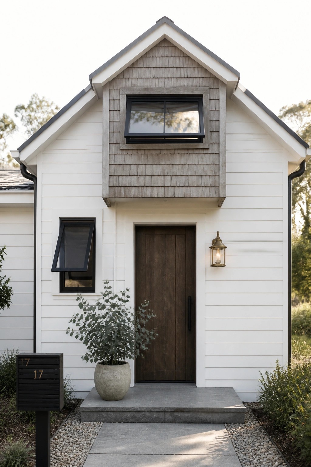 A front view of a modern farmhouse with white horizontal siding, a greige wood shingle gable above the entrance, a dark wooden door, and black-framed windows.
