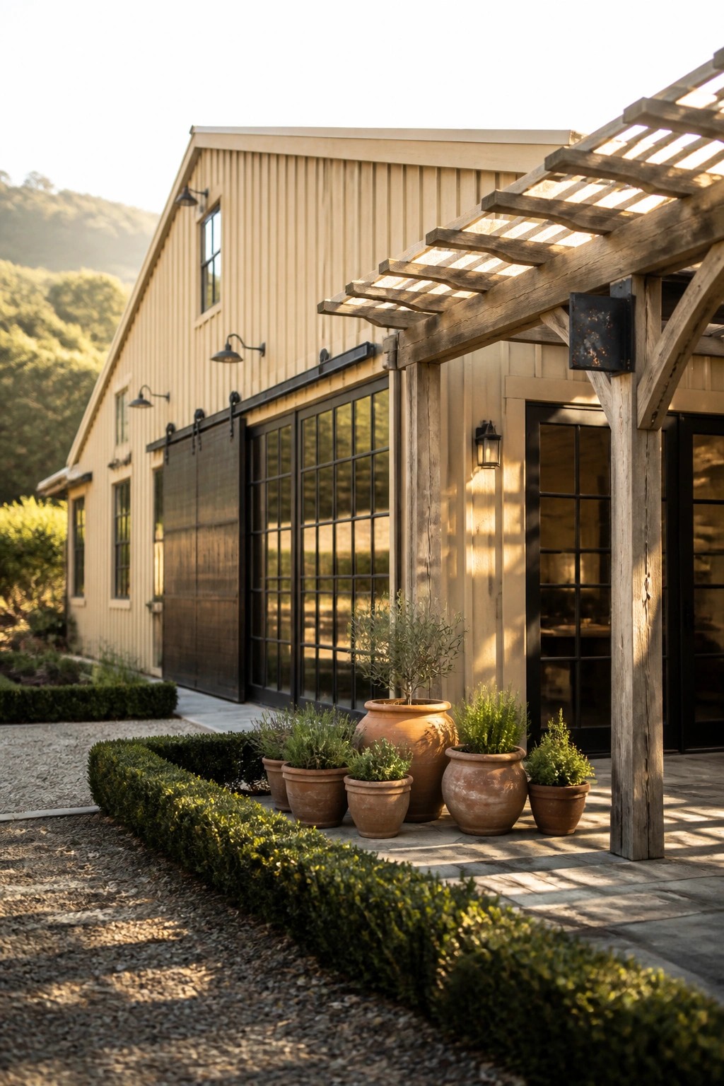 Beige barn-style building with black doors, wooden pergola, and terracotta plant pots.