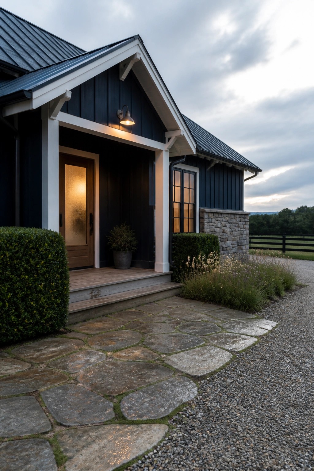 A house with dark navy siding, white trim, a stone foundation, a wooden front door, and a flagstone path beside a gravel driveway.