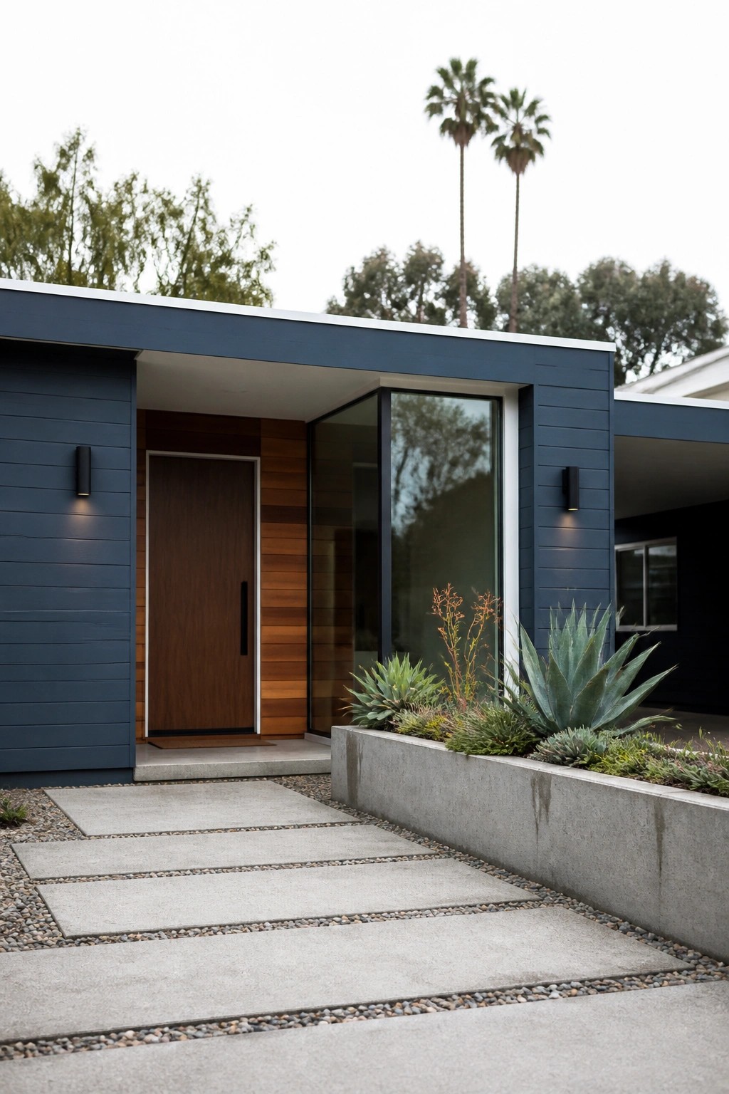Modern single-story house exterior with dark blue horizontal siding, wood paneling around the front door, and a concrete path bordered by gravel.