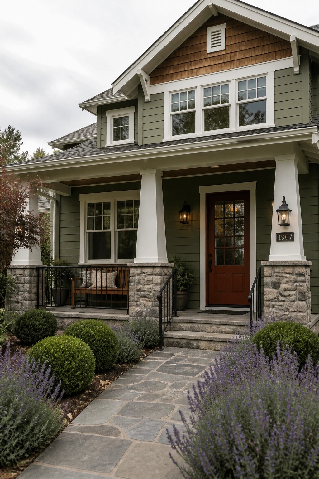 Front view of a two-story house with sage green siding, white trim, a red door, stone porch columns, and lavender landscaping along a stone walkway.
