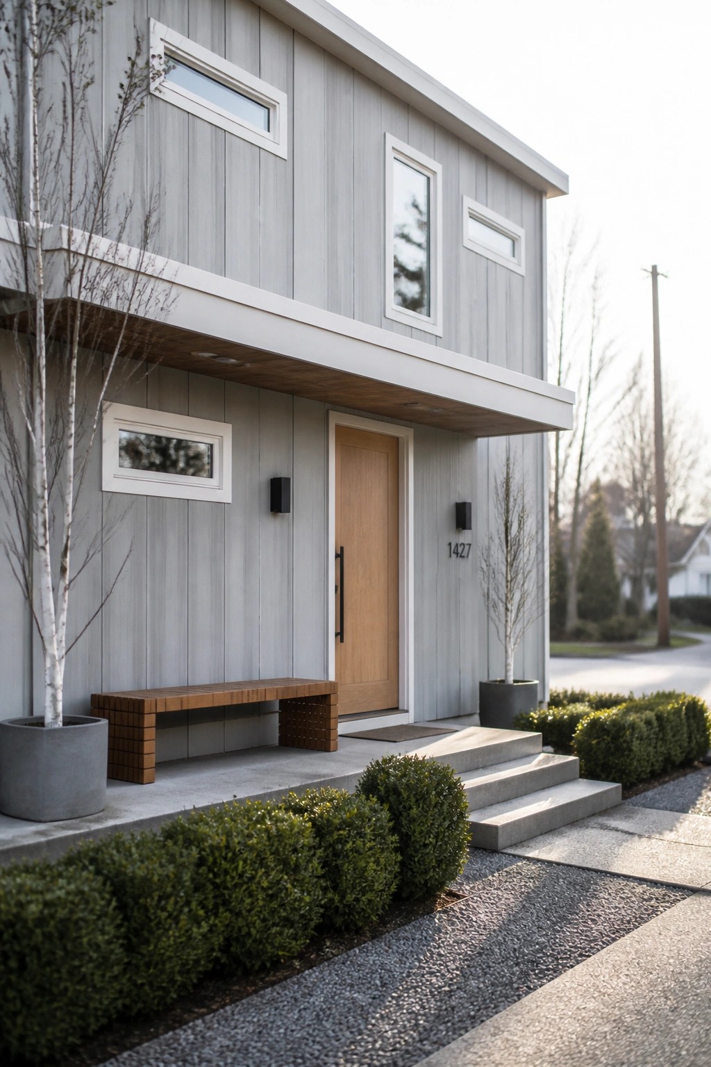 A modern two-story house with light gray vertical siding, white window trim, a light wood front door, and concrete steps leading to the entry.