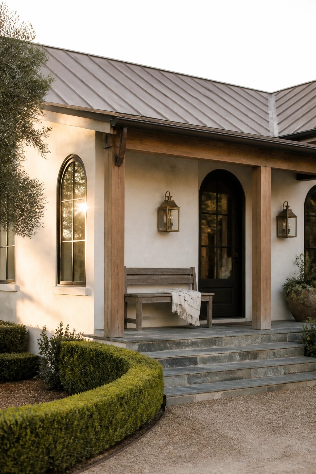 Front view of a white stucco house with wooden porch beams, a wooden bench on the porch, arched windows, a black door, and stone steps.