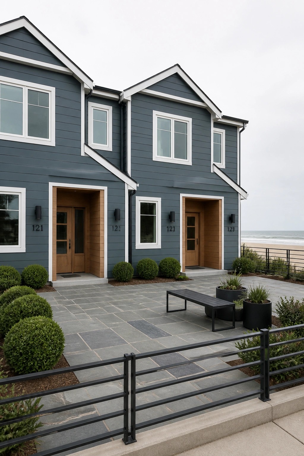 Two attached two-story homes with blue-gray siding, white trim, and wood entry doors on a stone patio facing the ocean.