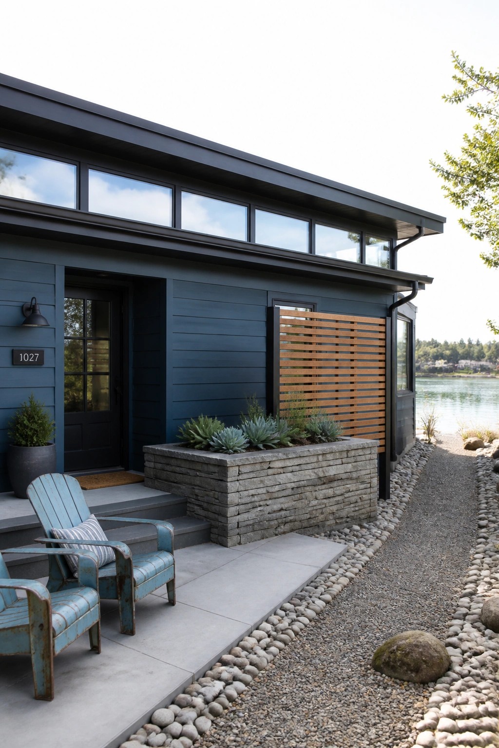 Modern house with deep blue siding, dark trim, stone planter box, and two weathered blue chairs on a concrete porch beside a gravel path and water.
