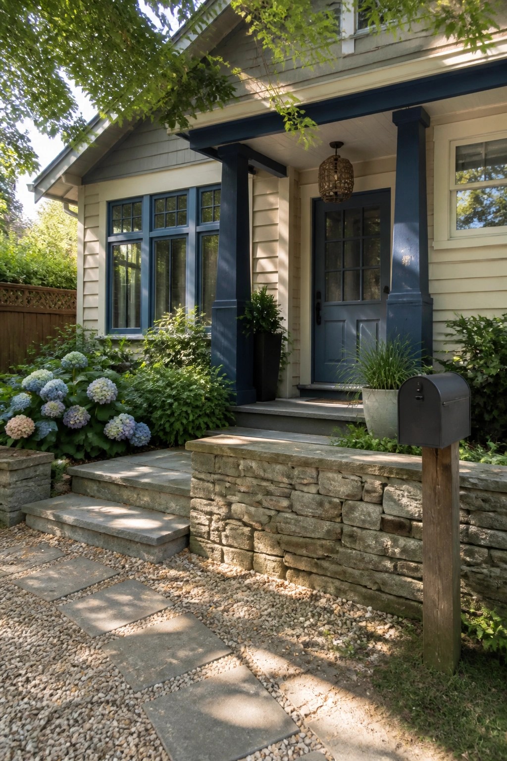 A light beige house exterior with dark blue trim, a blue front door, stone steps and retaining wall, and a gravel path bordered by hydrangeas.