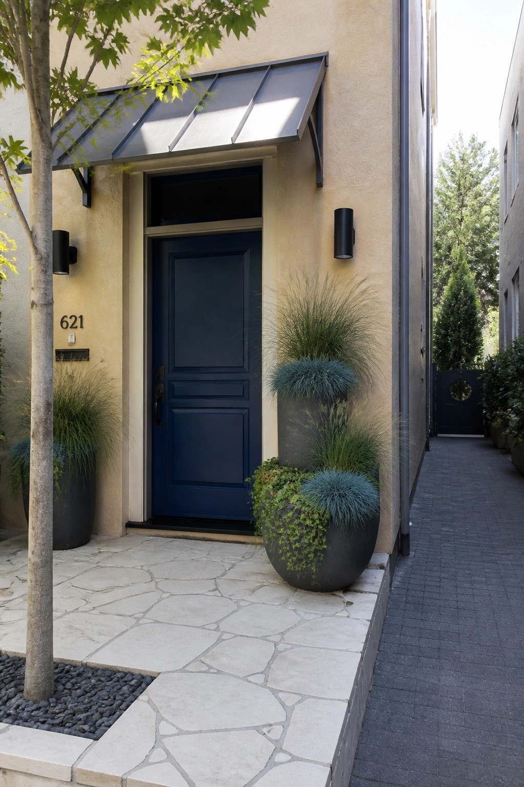 Navy blue door under black awning beside large planters with grasses on stone porch.