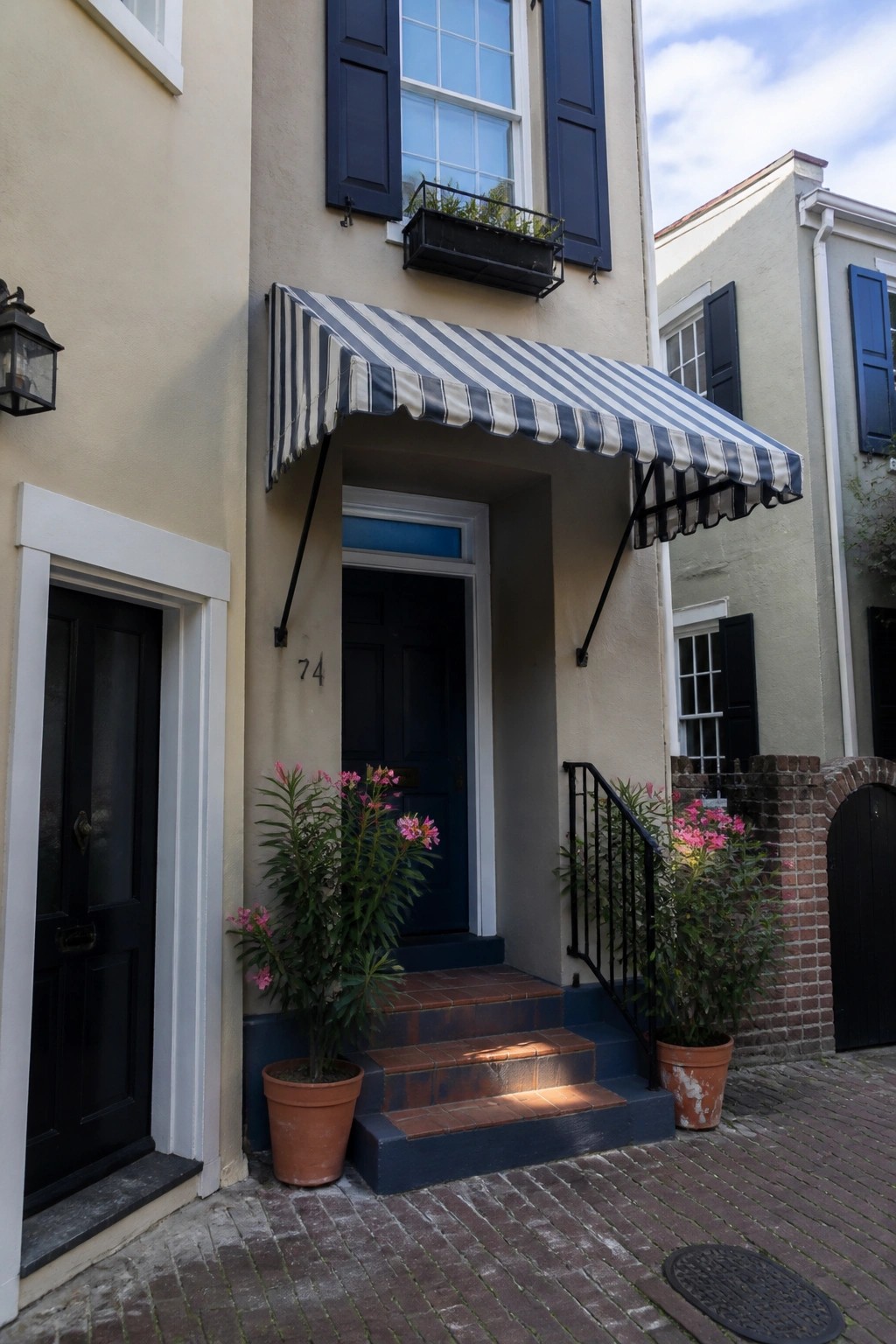 A beige house with a blue front door, striped awning above it, brick steps, and potted plants on either side.