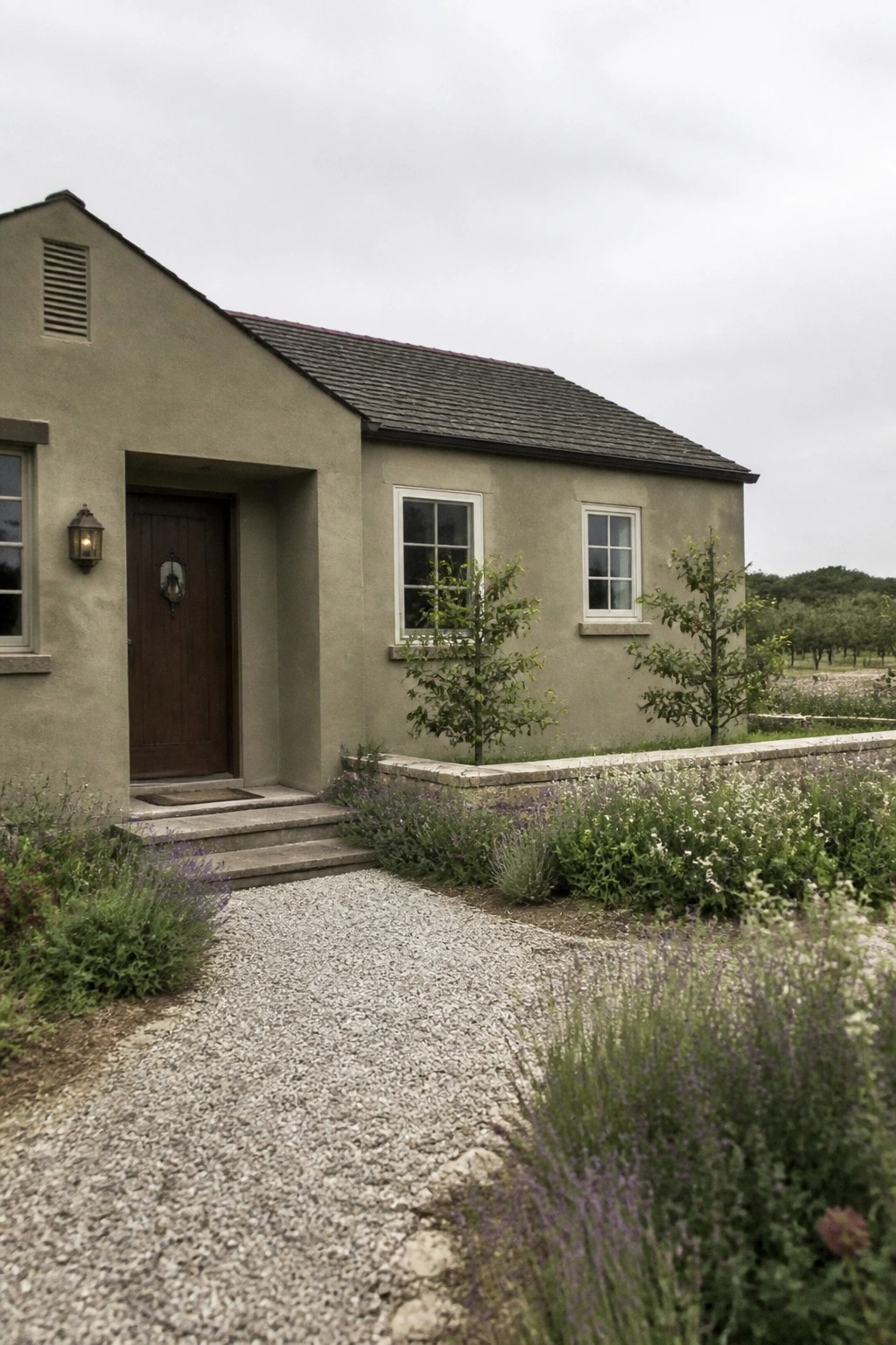 A rural house exterior painted in muted sage green stucco with a dark roof and gravel path.