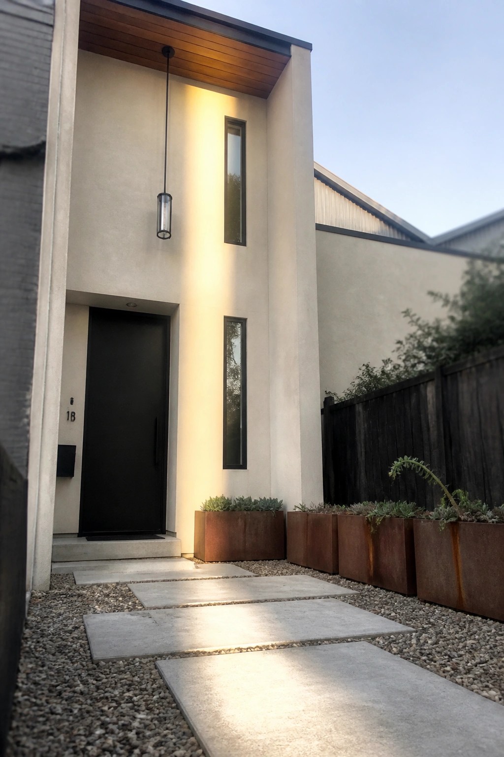 A light beige modern house exterior features a black front door, a hanging cylindrical pendant light, and two narrow vertical windows above a gravel path with concrete stepping stones and rusted metal planters.