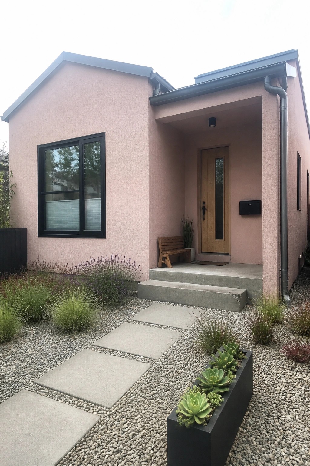A small light pink stucco house with a wooden front door, black-framed window, concrete steps, and gravel path with pavers.