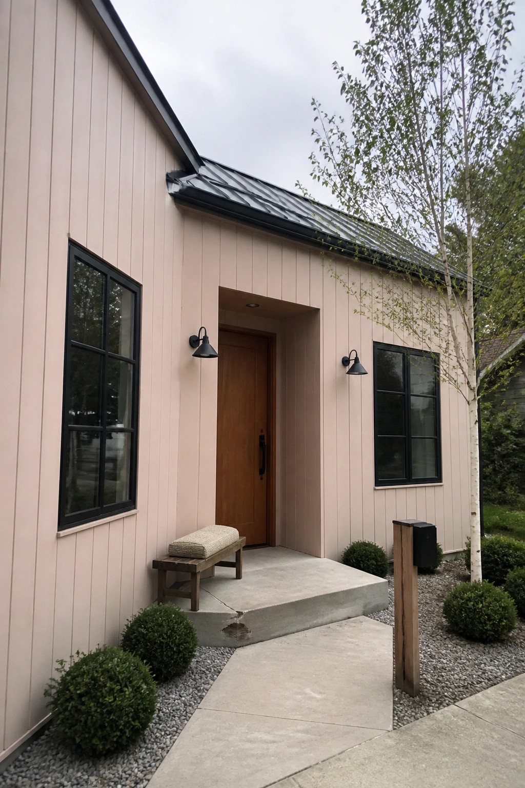 A small house with light pink vertical siding, black-framed windows, a wooden front door, and a concrete entry porch with two black sconces.