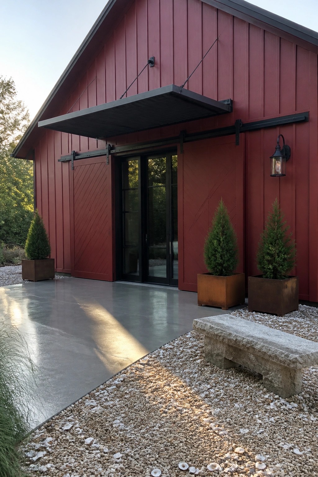 Red barn-style house exterior with black sliding barn doors, a flat black metal awning above the entrance, two potted evergreen trees in rusted metal planters, and a gravel area with a stone bench.