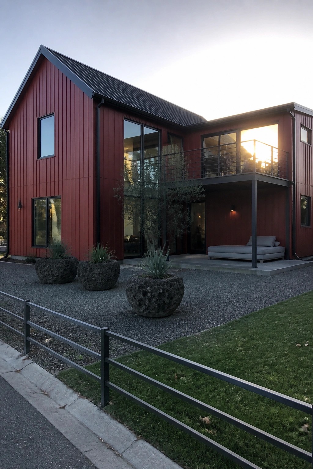 Modern red house with large windows, balcony, stone planters on gravel at sunset.