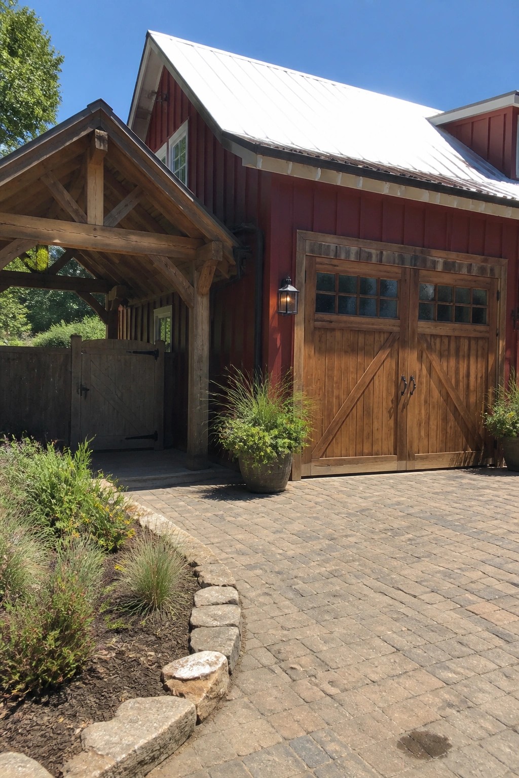 Red farmhouse exterior with natural wood double garage doors, wooden overhang beams, cobblestone driveway, and potted plants along the edge.