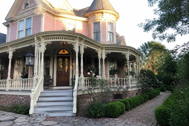 A pink Victorian house with a conical turret, wrap-around porch with white ornate trim, wooden front door, and cobblestone path.