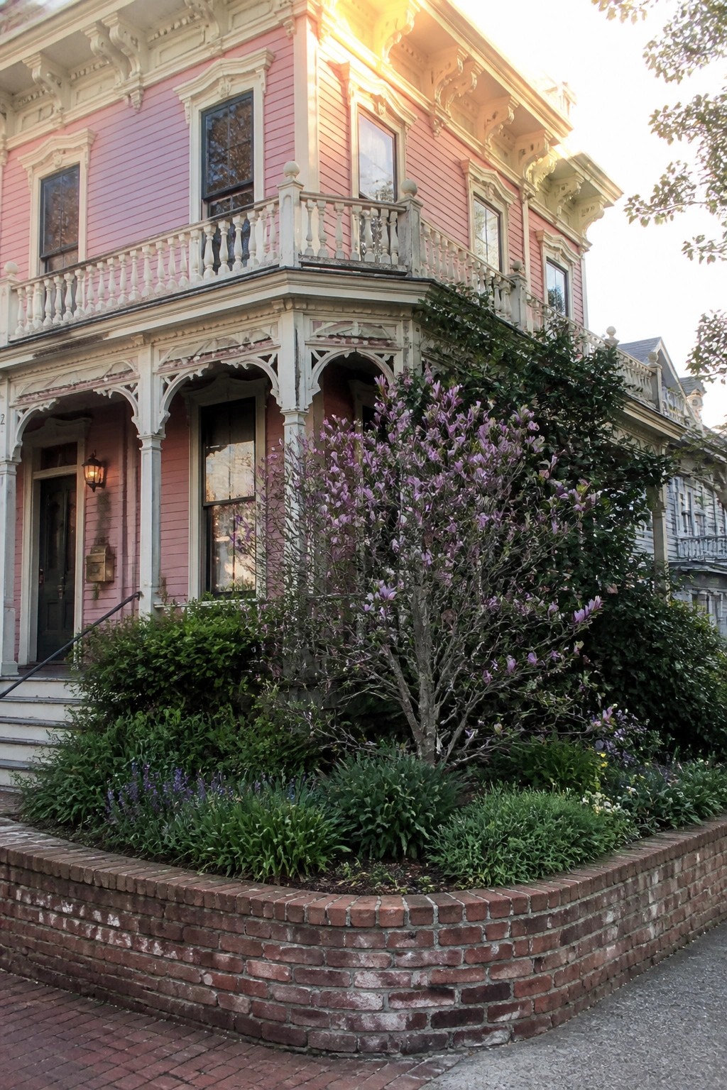 Pink Victorian house with white ornate trim, second-floor balcony, and brick-edged garden bed with flowering shrubs in front.