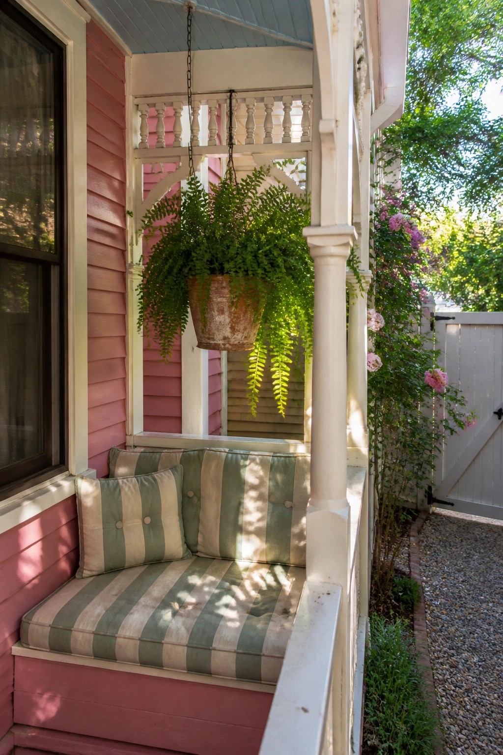 A pink wooden house exterior with white trim shows a built-in striped bench on the porch and a hanging fern in a terracotta pot.