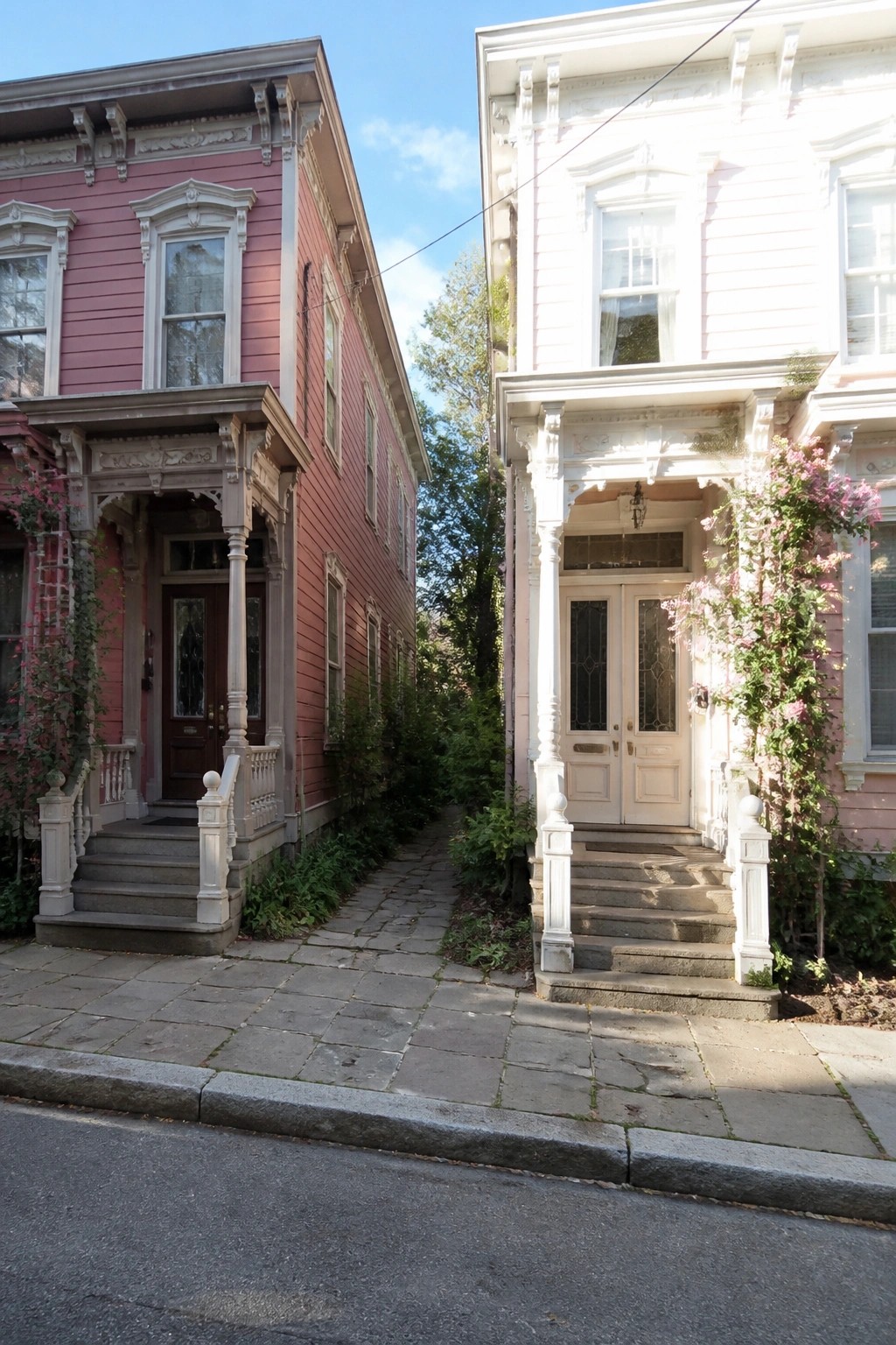Two adjacent Victorian houses side by side on a city street, one painted deep pink and the other pale pink, both featuring white ornate trim, covered porches, and stone steps.
