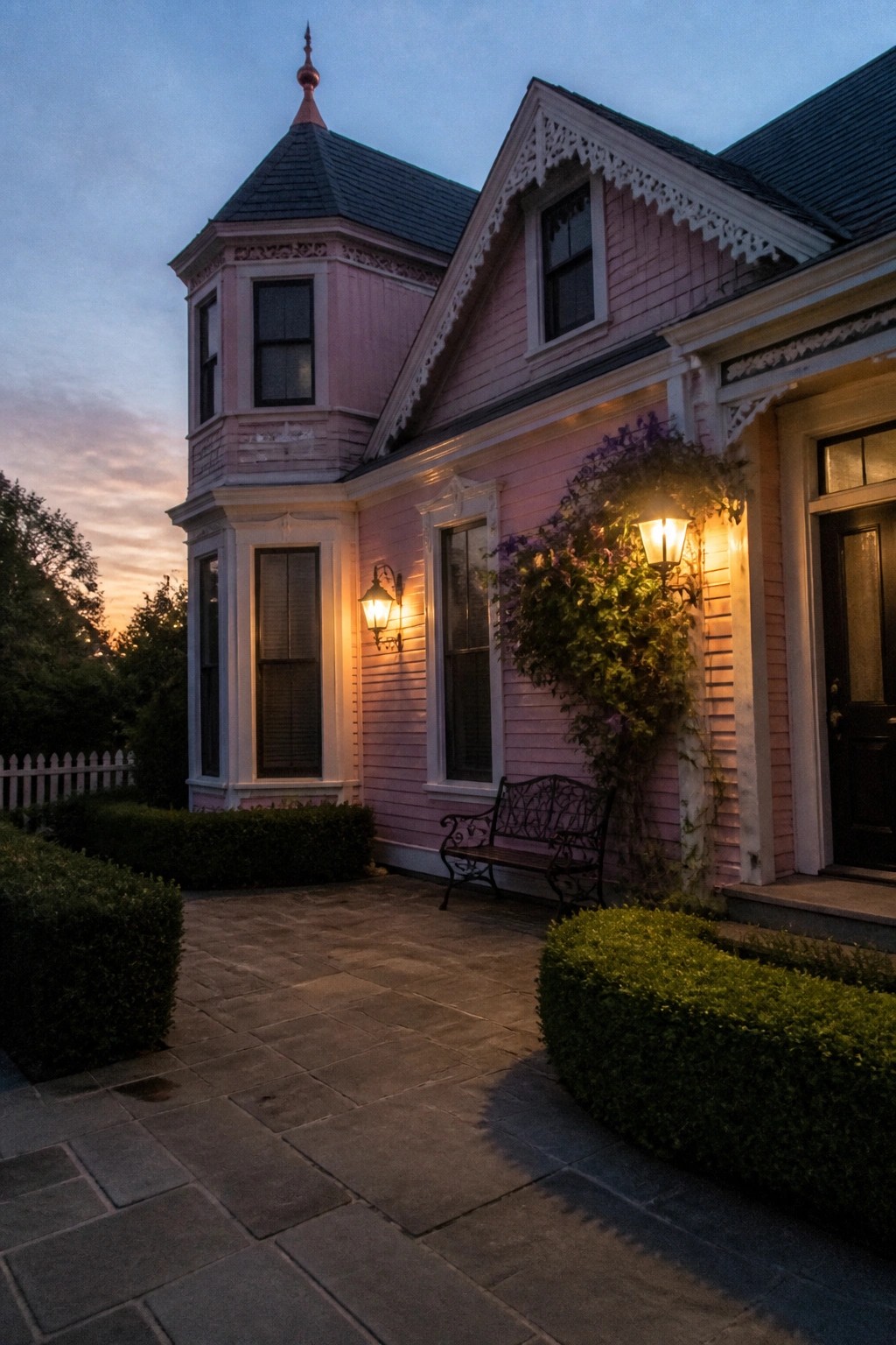 Pink Victorian house with lit lanterns, bench, and stone patio at dusk