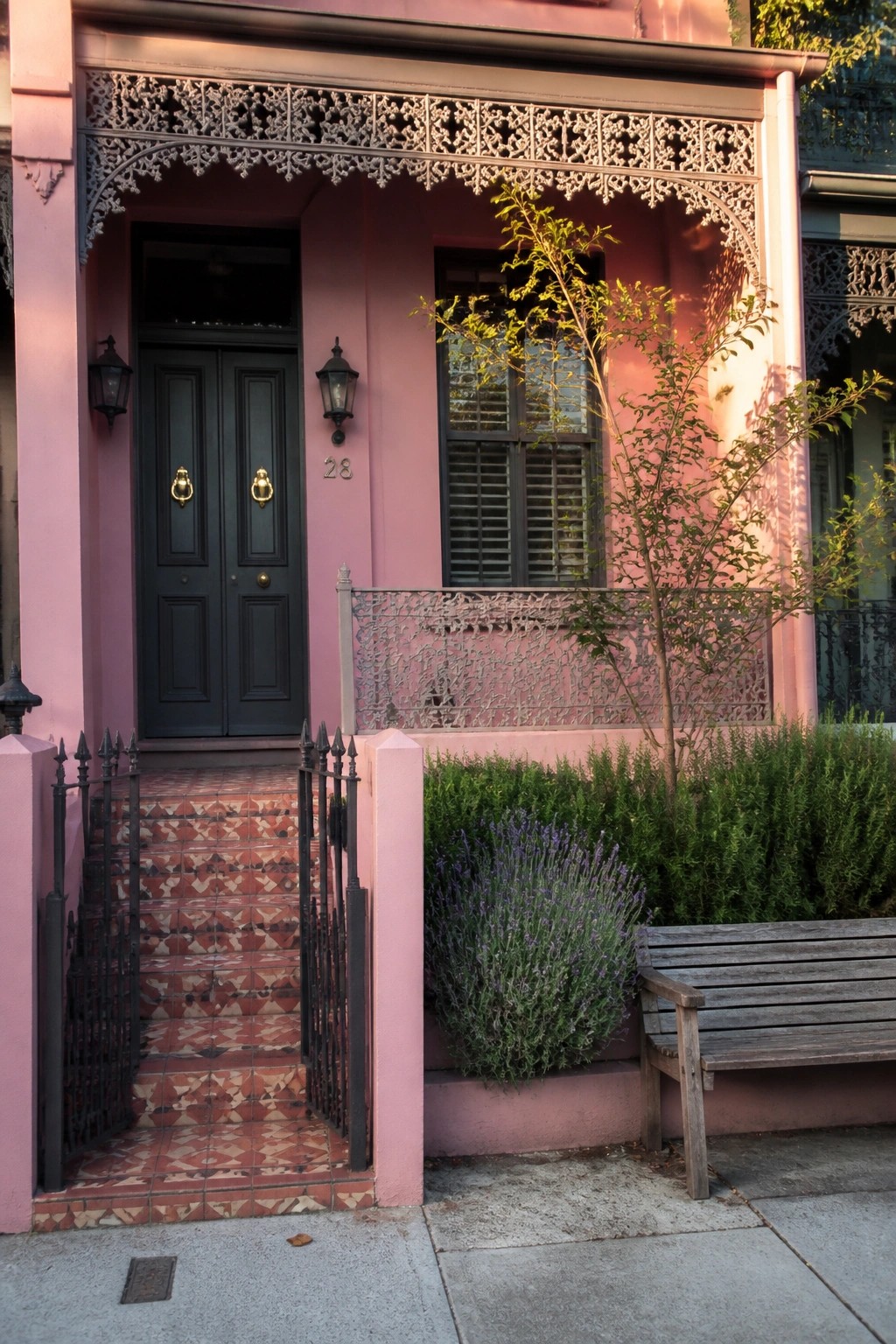 A pink Victorian house front with a black double door, gold hardware, ornate metal trim overhead, tiled steps, and a small garden area with a bench and lavender plants.