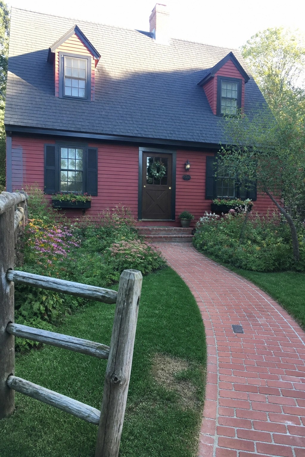 Red house with black shutters, brick path curving past wooden fence and garden.