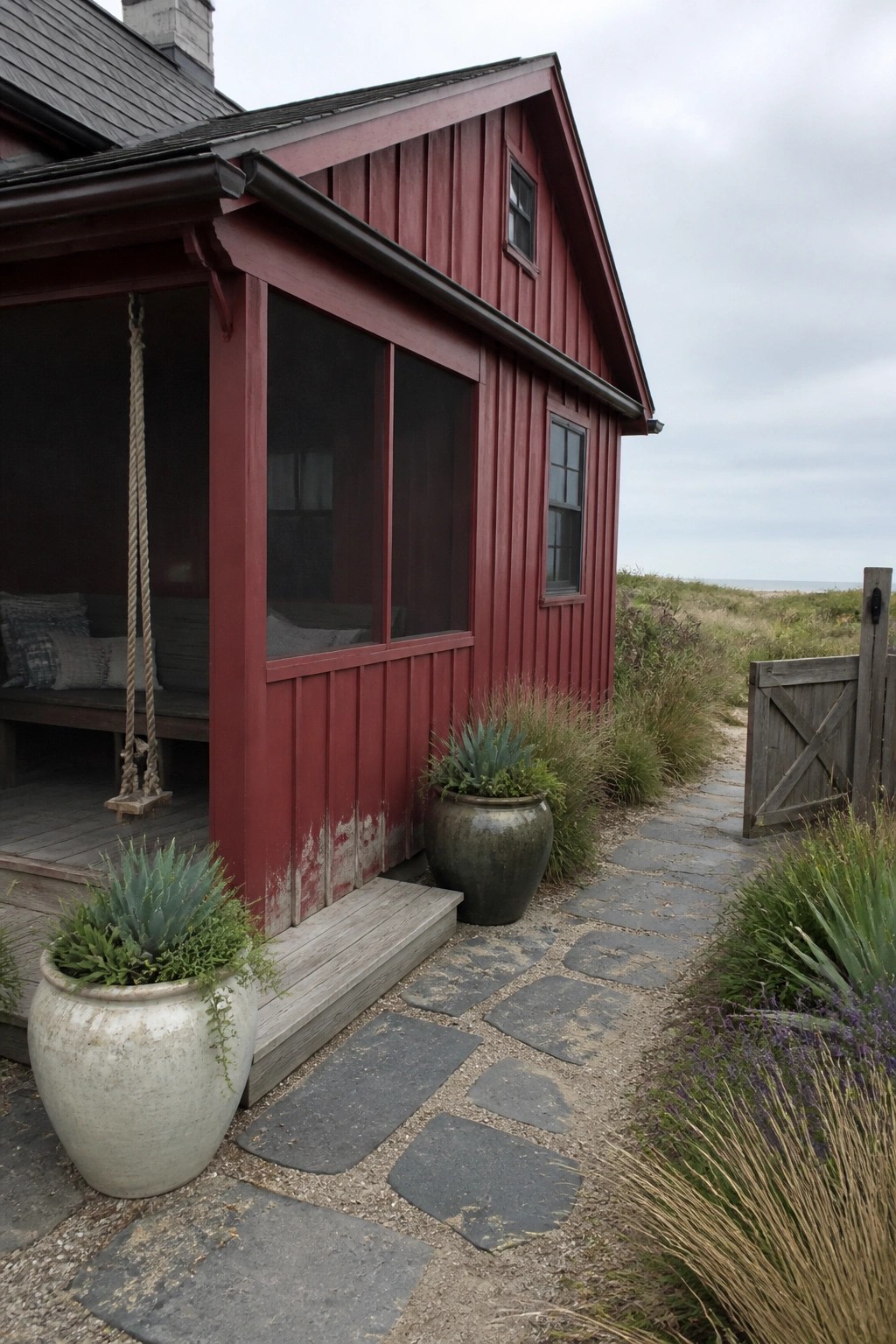 A red wooden house exterior with dark trim, a screened porch, potted plants, and a stone path beside a wooden gate.