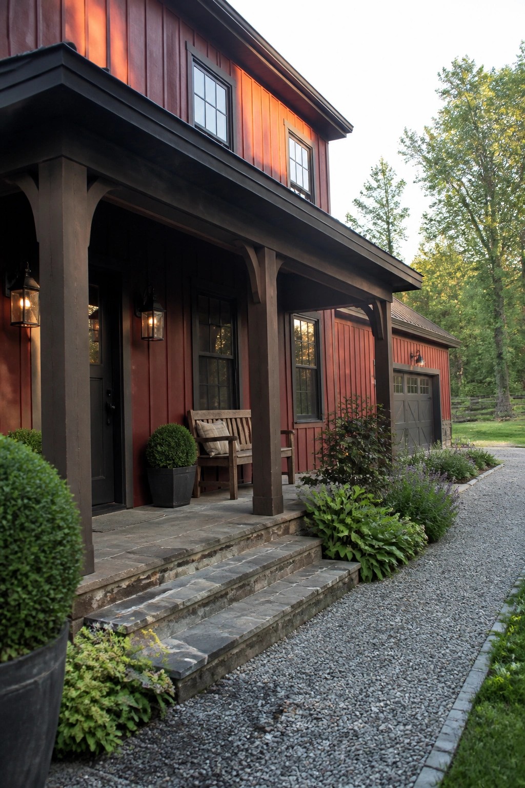 A red wooden house with dark trim, a charcoal roof, a covered porch with stone steps, and a gravel driveway beside landscaping.