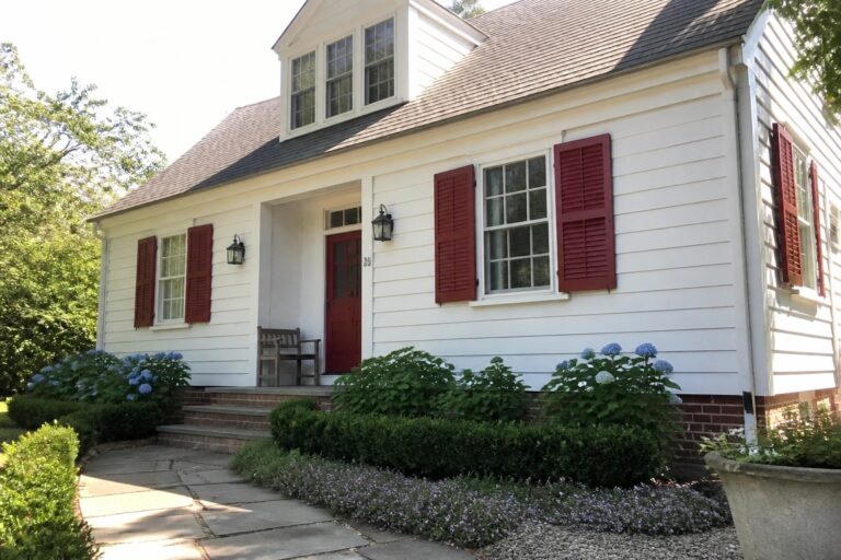 A white house with red shutters and a red front door, shown from the front with a stone path and garden beds.