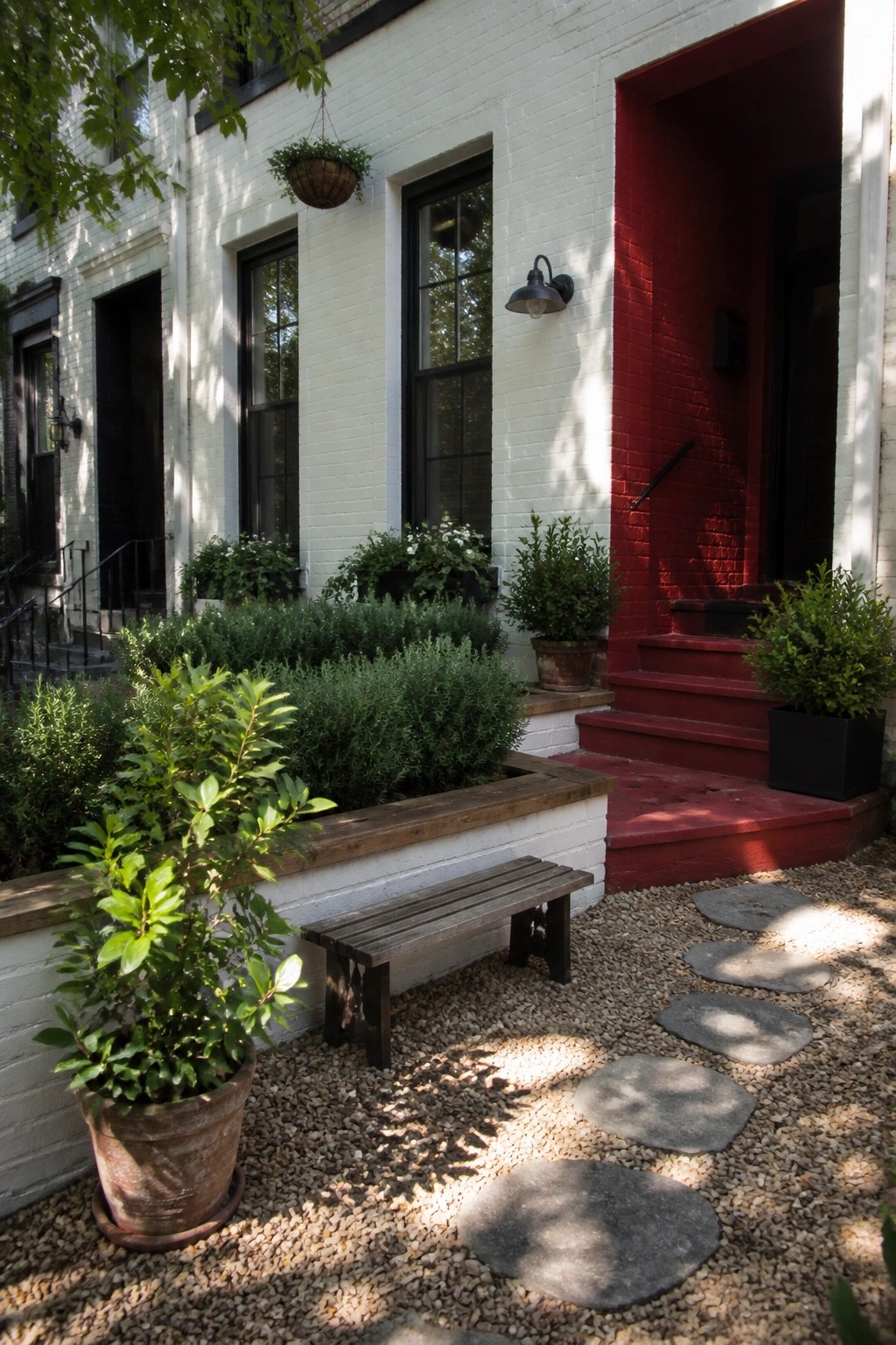 A white brick house exterior with a bright red door, black-framed windows, red steps, and potted plants beside a gravel path.
