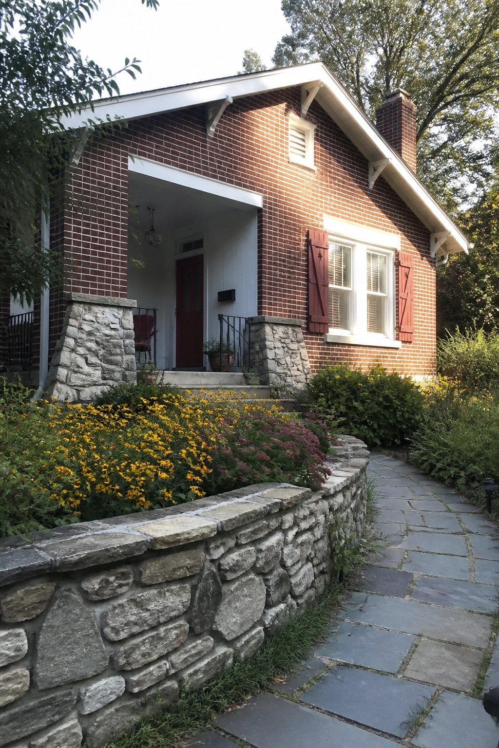 A red brick house exterior with white trim, a red front door, red shutters on one window, and a stone retaining wall along a paved path.